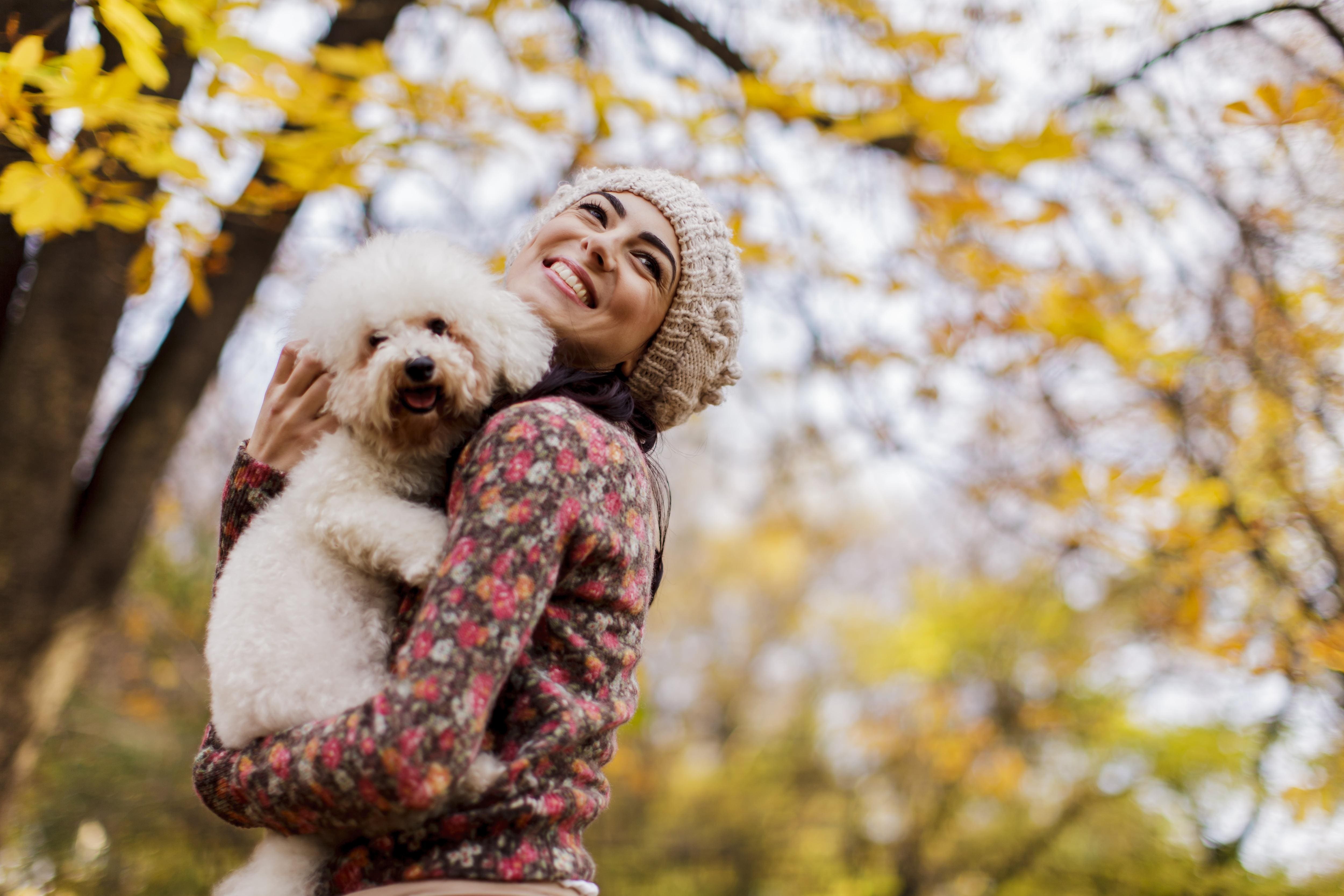 Brunette woman in knitted beanie and flowered shirt holds a shaggy dog with fall trees in background.